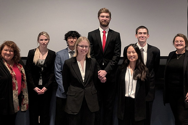 Group of eight people in business attire standing in two rows in front of a projection screen.